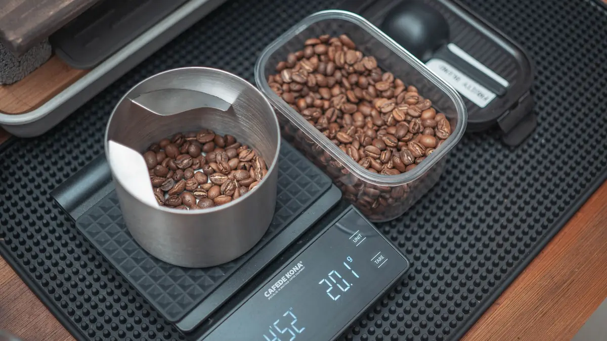 Des grains de café pesés dans un récipient posé sur une balance numérique, à côté d'une boîte de grains
