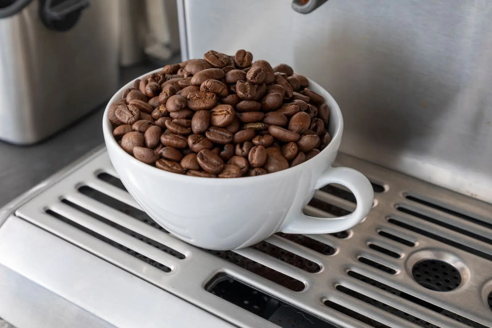 Une tasse blanche remplie de grains de café posée sur la grille d'égouttage d'une machine expresso : le grain avant la tasse