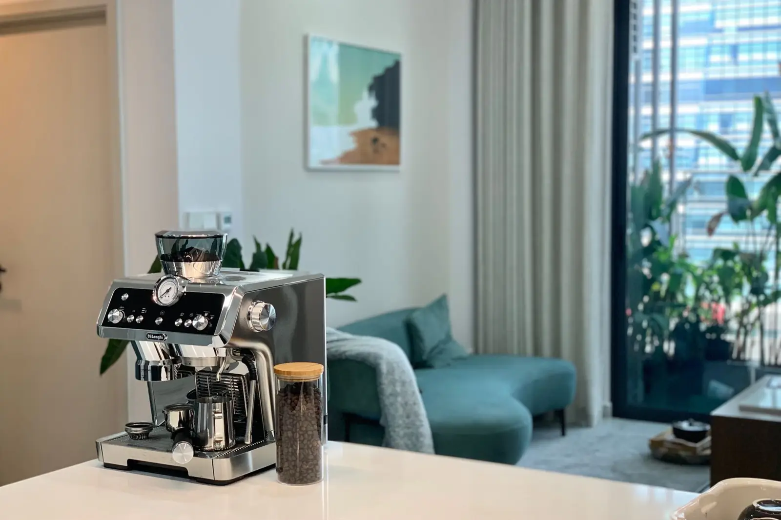 Une machine à café posée sur le plan de travail d'une pièce de vie lumineuse, un canapé et des plantes à l'arrière-plan