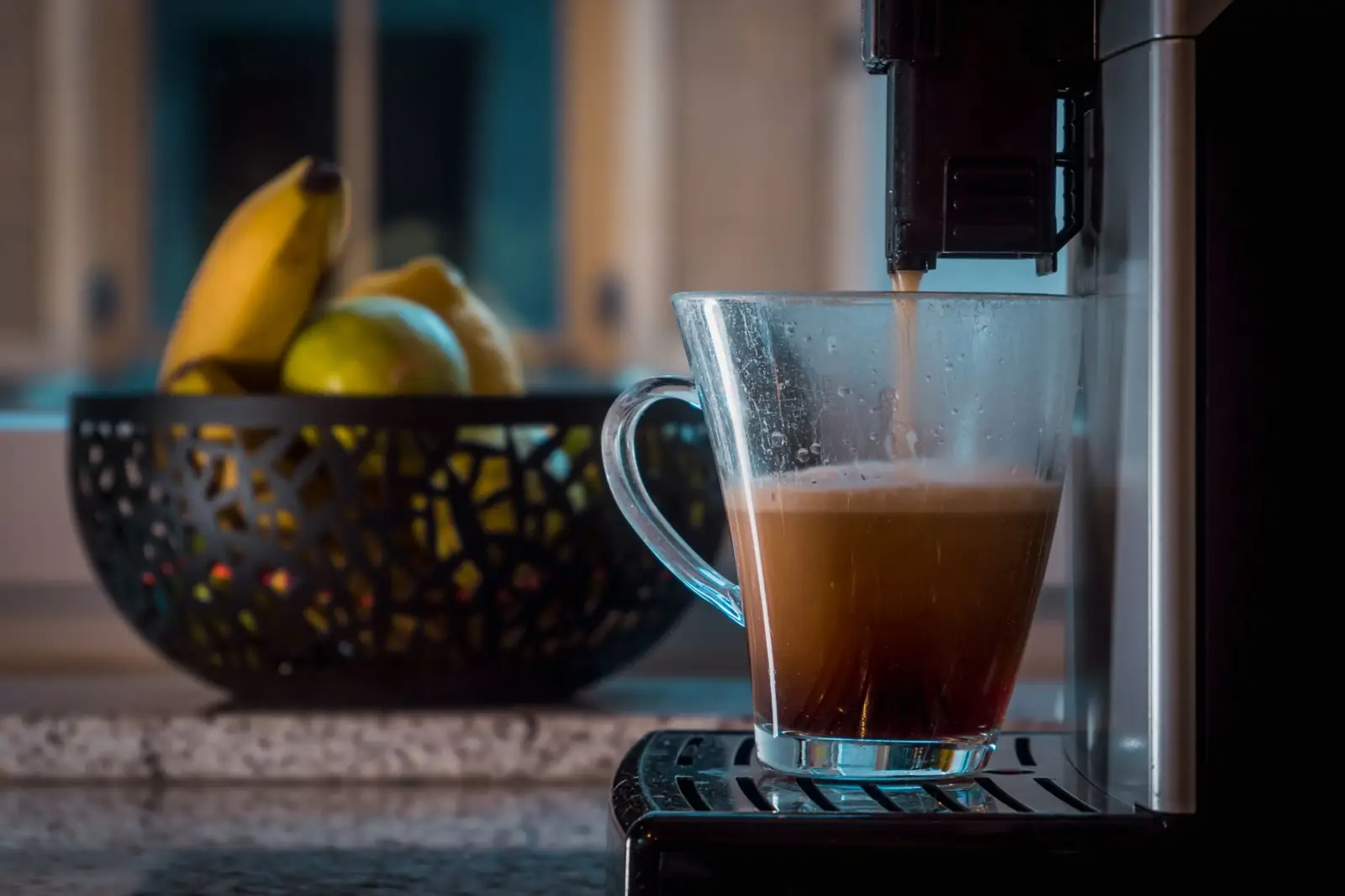 Un café coule d'une machine automatique dans une tasse en verre transparente, une corbeille de fruits à l'arrière-plan